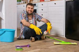 Caucasian young bearded man sits on the floor of his kitchen and tries to clean it using all of his detergents, rags, washcloths and brushes photo