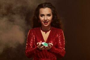 Young pretty lucky brunette woman in a red evening dress holds poker chips in her hands, showing her winnings. photo