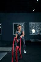 Vertical full length shot of an athletic fitness woman doing functional training exercise with battle ropes at the gym Cross Fit cross training strength endurance energy power concept photo