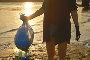 Young volunteer in black gloves is walking with garbage bag along a dirty beach of the river and cleaning up trash. People and ecology. Close-up. photo