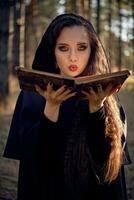 Witch in black, long dress with cape and hood. Posing in pine forest. Holding magic book and blowing on it. Spells, magic and witchcraft. Close-up. photo