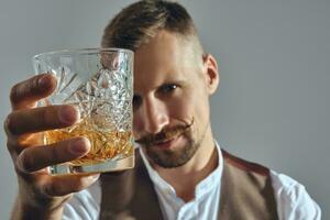 hombre con elegante Bigote, vestido en clásico marrón chaleco, blanco camisa es sentado a el mesa, disfrutando whisky. gris fondo, de cerca disparo. foto