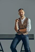 hombre con un elegante Bigote, vestido en marrón chaleco, blanco camisa y oscuro pantalones es sentado en mesa, disfrutando cigarro. gris fondo, de cerca. foto