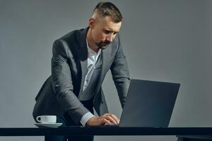 Bearded businessman with stylish mustache, dressed in a classic suit is working at laptop while standing at table in office, gray background. photo