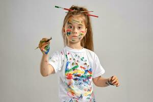 Little girl in white t-shirt, with brushes in her hair is posing standing isolated on white, gesticulating with painted hands and face. Close-up. photo