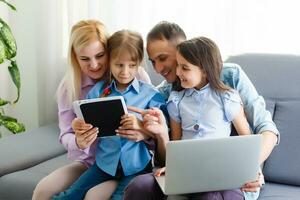 Happy family of four parents and cute little kids children enjoy using the tablet, watching cartoons, make internet video call or shopping online looking at computer screen sit together at home photo