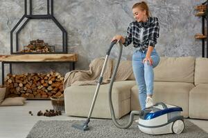 Young smiling woman vacuum cleaning the carpet in the living room, modern scandinavian interior. Home, housekeeping concept photo