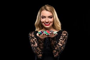 Elegant blonde in a black dress, casino player holding a handful of chips on black background photo
