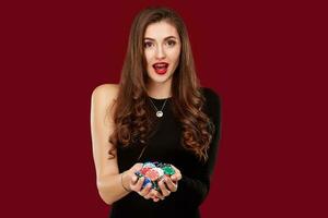 Casino, gambling, poker, people and entertainment concept - woman poker player in black dress with chips in hands on red background. photo