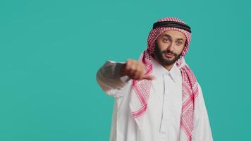 Muslim guy with ancient gown and kufiyah gives thumbs down and feels unsatisfied with stuff. Young person expresses an apparent denial and negative sign in studio, arabic model. video