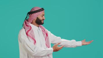 Muslim guy wearing traditional robe and ghutra gesturing to side with hands and posing for studio shot. Arabic smiling person promoting product while showing marketing ad aside. video