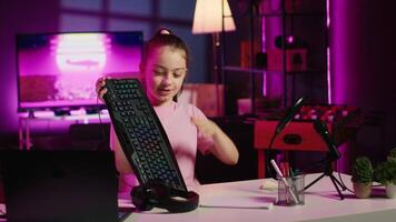 mignonne fille dans studio en utilisant caméra à la revue jeu clavier, Souris et écouteurs. joyeux enfant spectacle hôte tournage électronique transporter, en présentant câblé ordinateur périphériques à les spectateurs video