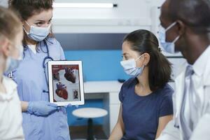 Detailed image of healthcare professionals using a tablet to explain to the female patients about the human heart. Nurse holds a device while the male doctor describes importance of medical checkups. photo