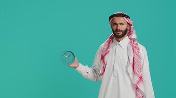 Arab guy sees wall clock to check time on camera, using timepiece to look at hour and minutes. Young person in traditional muslim clothing examining local timezone, standing over background. video