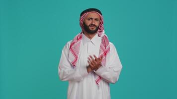 Arab adult dressed in traditional attire clapping hands in studio, congratulating and cheering on camera with confident expression. Muslim guy showing support and applauding people. video