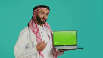 Islamic guy presents greenscreen on pc with smart isolated display for copyspace layout. Wearing thobe and headscarf, young person shows blank chromakey mockup on wireless computer. video