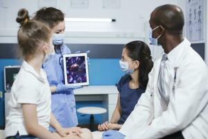 In the clinic, female nurse grasps a digital tablet showing a bacteria image in front of the patients and African American doctor. Expert care and education ensure healthcare and wellness. photo