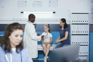 African American doctor examines a young patient, explaining the condition while wearing a lab coat. Effective communication promotes health in this calm and professional setting. Focus on background. photo