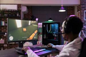 African american man in brick wall apartment playing video games on gaming PC at computer desk, enjoying day off from work. Gamer battling enemies in online multiplayer shooter photo