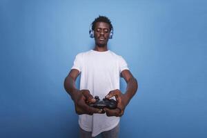 African American video gamer in studio using controller and headphones. Black man playing an internet game on a console with a joystick while standing over isolated blue background. photo