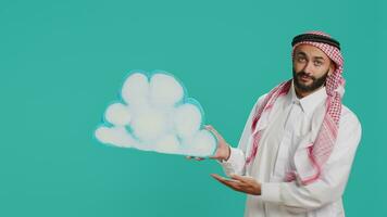 Middle eastern guy presents cloud board in studio, holding isolated billboard cutout in natural shape. Person wearing traditional arab islamic clothing and headscarf doing new web advertisement. video