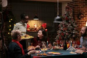 Cheerful people at festive dinner, meeting to celebrate christmas eve together with family. Diverse men and women serving meal and drinking wine glasses, feeling joyful during winter season. photo