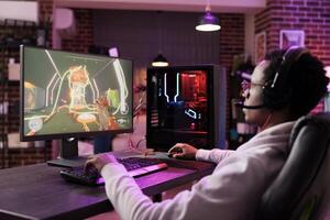 Man in brick wall living room playing engaging video games on gaming PC at computer desk, chilling after work. Gamer contending against foes in online multiplayer shooter photo