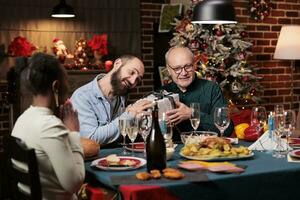 Two men exchanging gifts at christmas dinner, diverse people offering present to spread positivity near fireplace. Father and son feeling cheerful with boxes of products on xmas celebration. photo