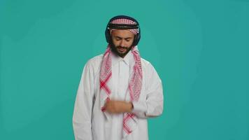 Joyful person dancing on songs in studio, listening to modern music on headphones and having fun on camera. Middle eastern adult in traditional clothing showing groovy dance moves. video