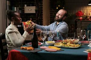 Diverse people exchange presents for christmas eve celebration, spreading positivity at festive dinner. Joyful man giving gift box to african american friend during december celebration. photo