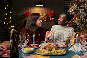 diverso mujer celebrando Navidad con cena, disfrutando estacional invierno evento a hogar. personas teniendo divertido con diciembre celebracion a mesa rodeado por festivo adornos y Navidad árbol. foto