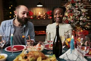 africano americano mujer a festivo cena celebrando diciembre fiesta con amigos reunión alrededor mesa y comiendo tradicional alimento. grupo de contento personas disfrutando Navidad víspera a hogar. foto