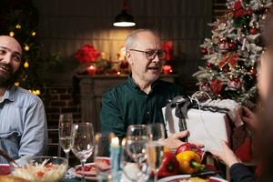 Senior people offering presents on xmas, spreading positivity and christmas spirit on winter holiday at home. Happy old couple celebrating seasonal december event with festive ornaments. photo