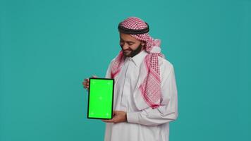 Arab person shows greenscreen display over studio background, presenting isolated chromakey mockup template. Young guy in islamic attire holding tablet with blank copyspace screen. video