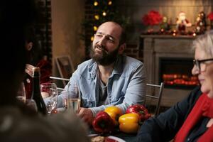 Happy persons talking about life and enjoying december holiday at home, grandparents and family members having fun eating traditional food and drinking wine. Diverse people gathering around table. photo