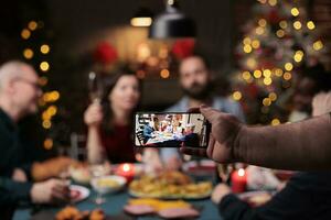 hombre tomando foto de familia a cena, celebrando Navidad víspera fiesta con tradicional comida y beber. alguien toma imágenes de personas disfrutando banquete con lentes de vino durante celebracion.