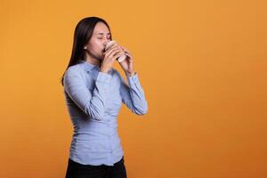 Filipino model holding cup of coffee drinking hot cappuccino enjoying free time before start to shoot in studio. Excited attractive woman with brunette hair standing over yellow background photo