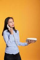 Filipino woman having remote conversation with friend, talking at landline phone in studio over yellow background. Cheerful young adult answering call discussing about business project photo