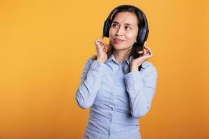 Cheerful woman with headset realaxing listening music, dancing over yellow background. Asian adult having fun in studio, showing dance moves during break time. Entertainment concept photo