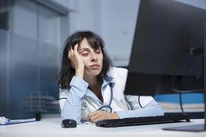 joven hembra médico vistiendo laboratorio Saco y estetoscopio, que cae dormido en frente de un computadora en clínica oficina. mirando cansado en un hospital habitación es un caucásico médico con mano en su rostro. foto