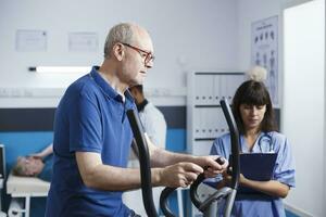 Elderly man is accompanied by female physician wearing blue scrubs as he receives physical therapy on stationary bike. Old patient is receiving rehabilitative care from medical practitioner. photo