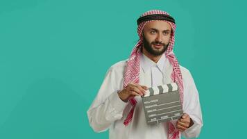 Middle eastern man holds clapperboard used for action takes on movie production, male producer using cinematography film slate. Muslim filmmaker wearing traditional arabic clothing. video