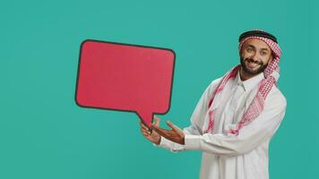 Arab person showing speech bubble in studio, creating advertisement for new commercial. Young adult working on new marketing slogan with isolated billboard icon, cardboard sign. video