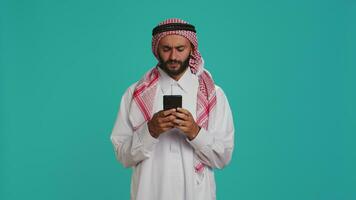 islamico uomo lavori su il suo mobile gadget, fare surf attraverso sociale media piattaforme o in linea pagine. mezzo orientale tipo nel consuetudine abbigliamento usi smartphone per accesso globale ragnatela, sensazione emozionato. video