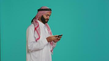 Muslim man wears traditional attire and playing videogames on smartphone, standing over blue background. Arab person enjoying mobile gaming competition on phone app, entertainment. video