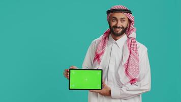 arabe homme montrant écran vert sur tablette plus de bleu arrière-plan, en présentant isolé fond maquette modèle. Jeune adulte dans islamique vêtements en portant dispositif avec Vide chromakey afficher. video