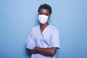Portrait of black man wearing face mask and uniform standing in studio, working for healthcare. Medical assistant looking at camera, having protection against coronavirus. photo
