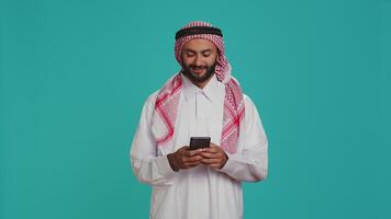 Muslim person in traditional costume having smartphone in hands and typing in internet social media messaging. Young adult browsing on mobile phone and texting, internet webpage. video