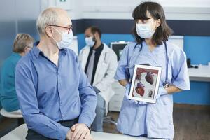 Female nurse in blue scrubs explains cardiology treatment displayed on device screen. Physician and elderly patient with face masks analyze image of heart on digital tablet. Woman with gadget. photo