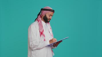 Arab person writing letter on files standing over blue background on camera, working on new schedule and taking notes. Guy in islamic clothes looking at checklist for paperwork in studio. video